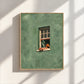 Sage green window artwork with a reader, displayed in a light wood-tone frame on a sunlit wall.