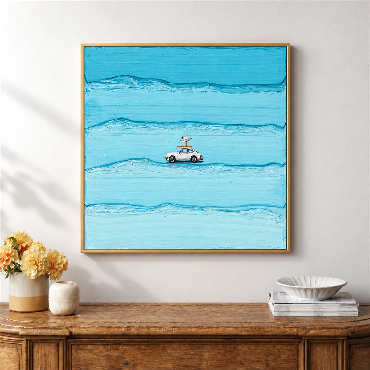 Blue car and surfer artwork in a light wood-tone frame above a rustic console with flowers and books.