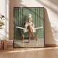 Bath reader painting in a brown wood frame leaning against a sunlit wall beside books.
