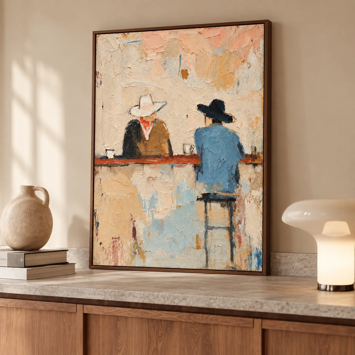 Cowboy bar painting in a brown wood frame resting on a sideboard beside a ceramic vase and lamp.