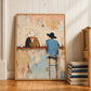 Western cowboy painting in a light wood frame leaning against white paneling beside a staircase and books.