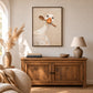 Spoon duck painting in a brown wood frame above a rustic sideboard with a lamp and vase.