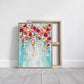 Bloom Cascade painting in a slim gold frame leaning on a pale floor in front of a reverse-facing canvas.