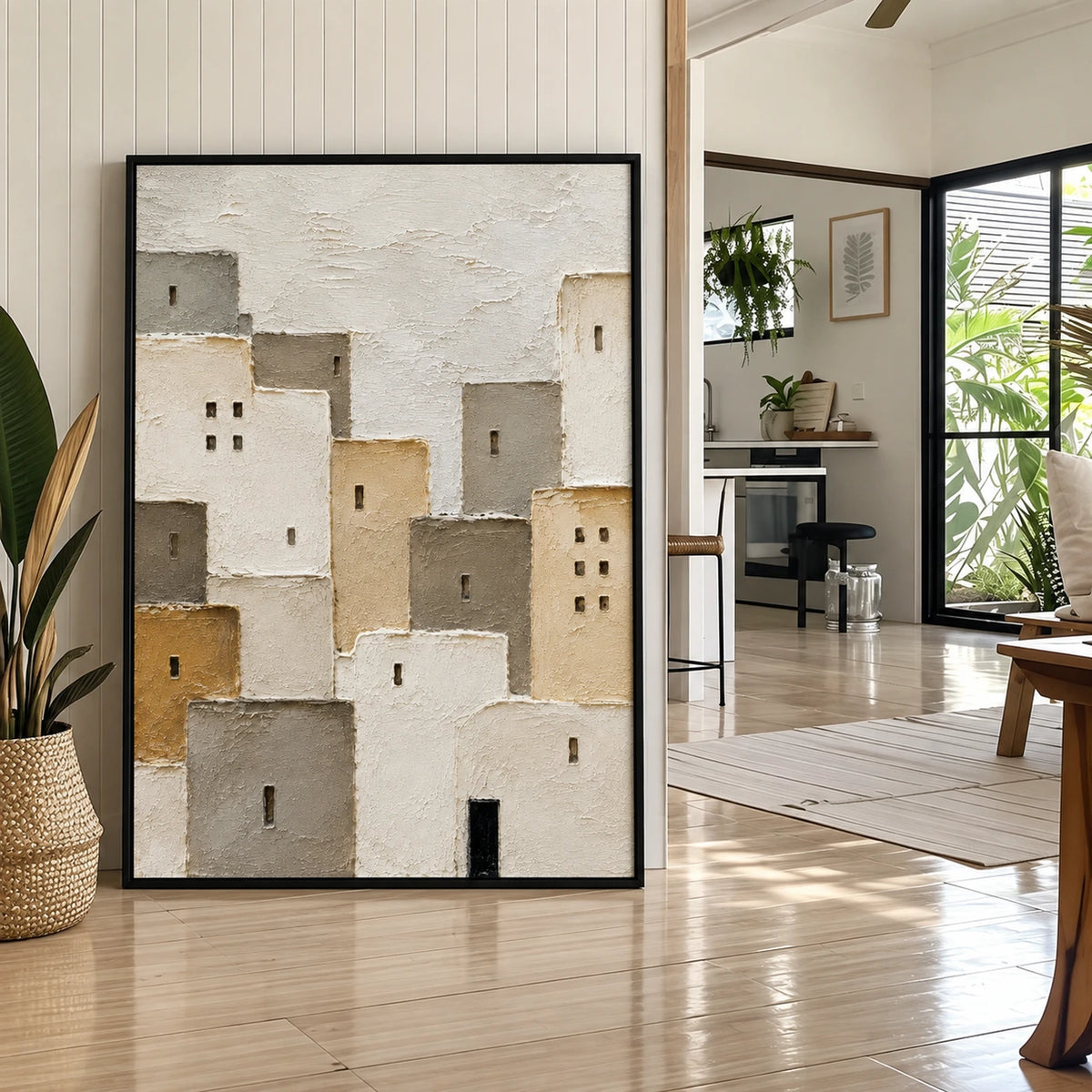Quiet Village painting in a black frame leaning against a paneled wall beside a large houseplant.