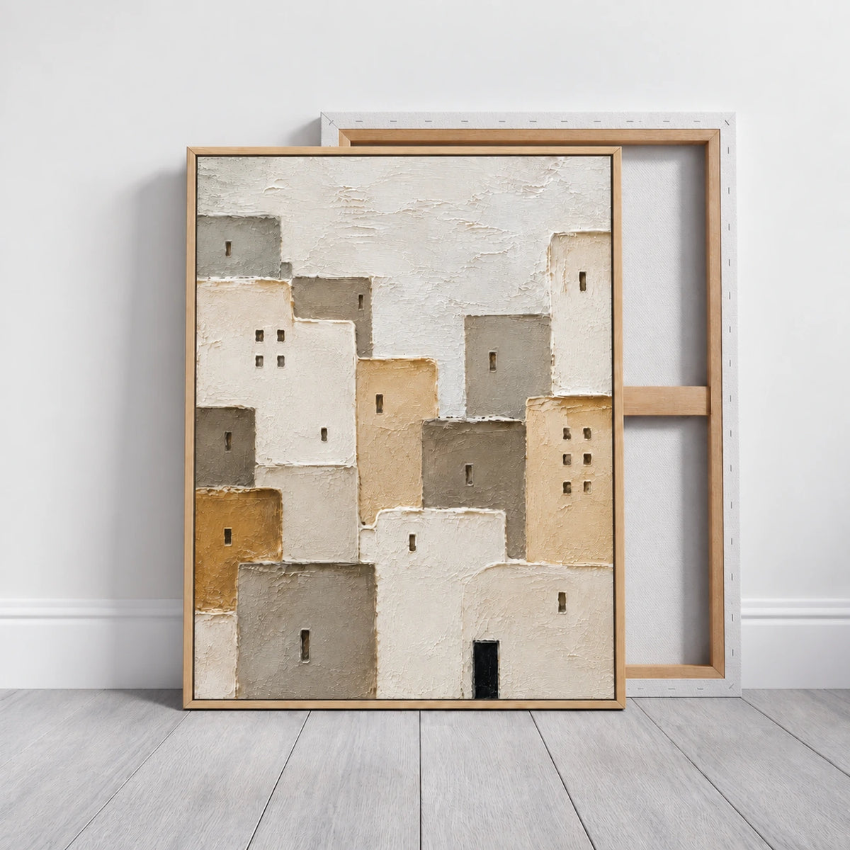 Neutral village painting in a light wood frame leaning in front of a reverse-facing canvas.