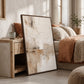 Framed Quiet Earth painting leaning beside a bedside cabinet and a bed with warm neutral linens.