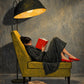 Full artwork of a reclining reader in dark clothing and red shoes, an olive armchair, a glowing black lamp and a stack of books.