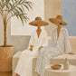 Full artwork of two seated straw-hatted figures in white and striped clothing beside a potted palm, pale arch and simple vessels.