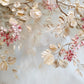 Trailing branches sweep across a misty blue-gray ground, carrying layered ivory leaves and clusters of soft pink blossoms. Full 2:1 artwork without a frame.