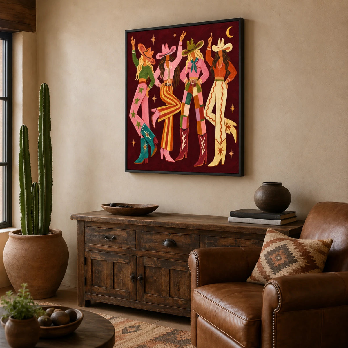 Cowgirl Revelry artwork displayed above a rustic wooden cabinet beside a brown leather armchair.