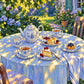 Sunlit Tea full artwork. A garden table set with patterned porcelain and small pastries catches patches of golden sunlight beneath surrounding foliage.
