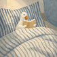 Bedtime Duck full artwork. A small white duck reads a book while tucked beneath blue-and-cream striped bedding.
